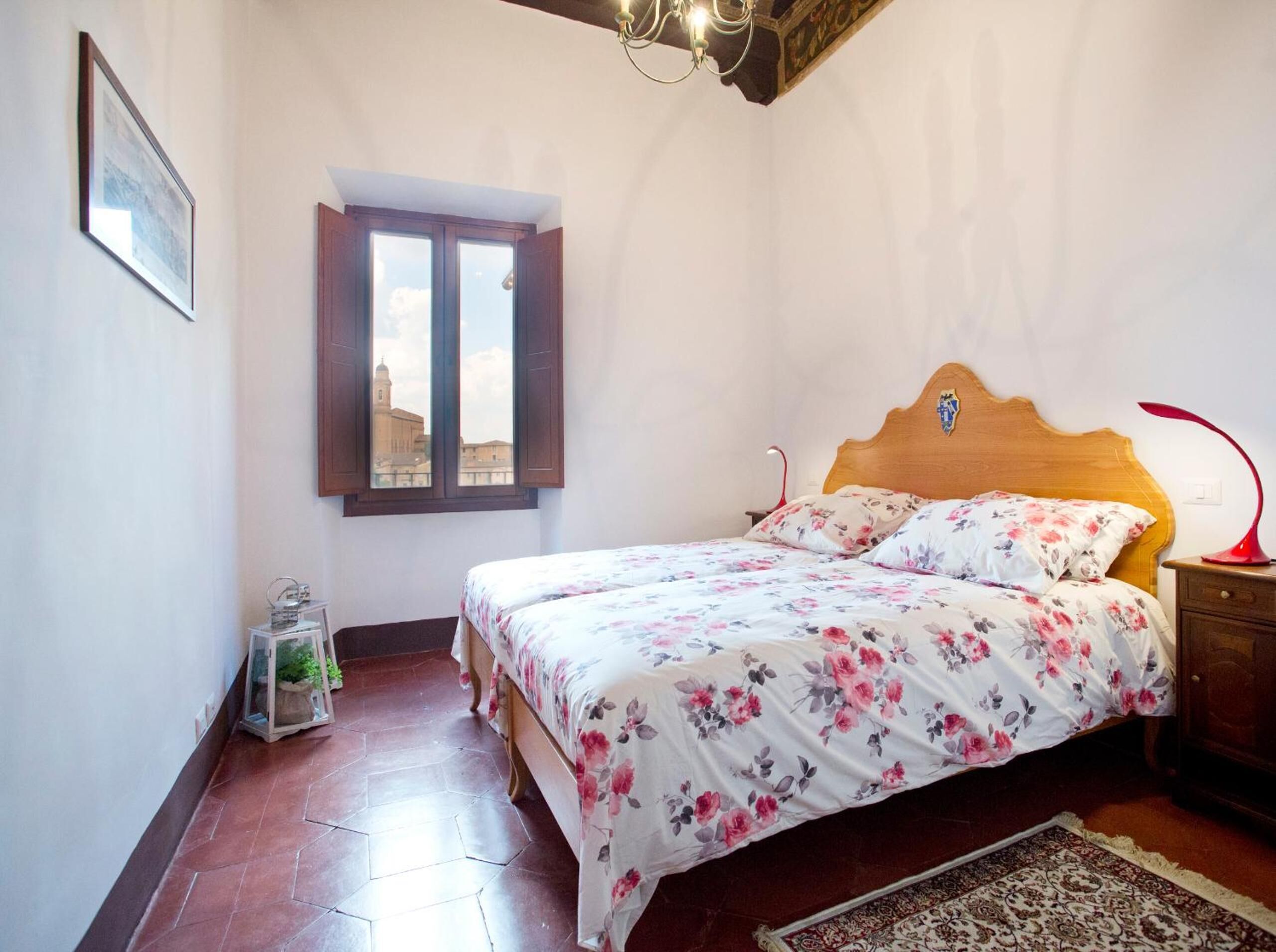 Een slaapkamer met een houten bed, bloemenbeddengoed, uitzicht door een raam en een rode lamp op een nachtkastje.
