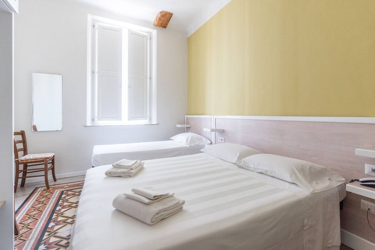 A hotel room with a large bed, a smaller bed, a chair, a window, and a bedside table.