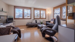 Een moderne woonkamer met een bank, fauteuil en salontafel. Grote ramen bieden uitzicht op besneeuwde bergen.