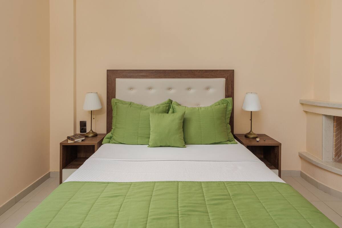 Een net opgemaakt bed met groene beddengoed, twee bedlampjes en een houten hoofdbord in een kamer met beige wanden.