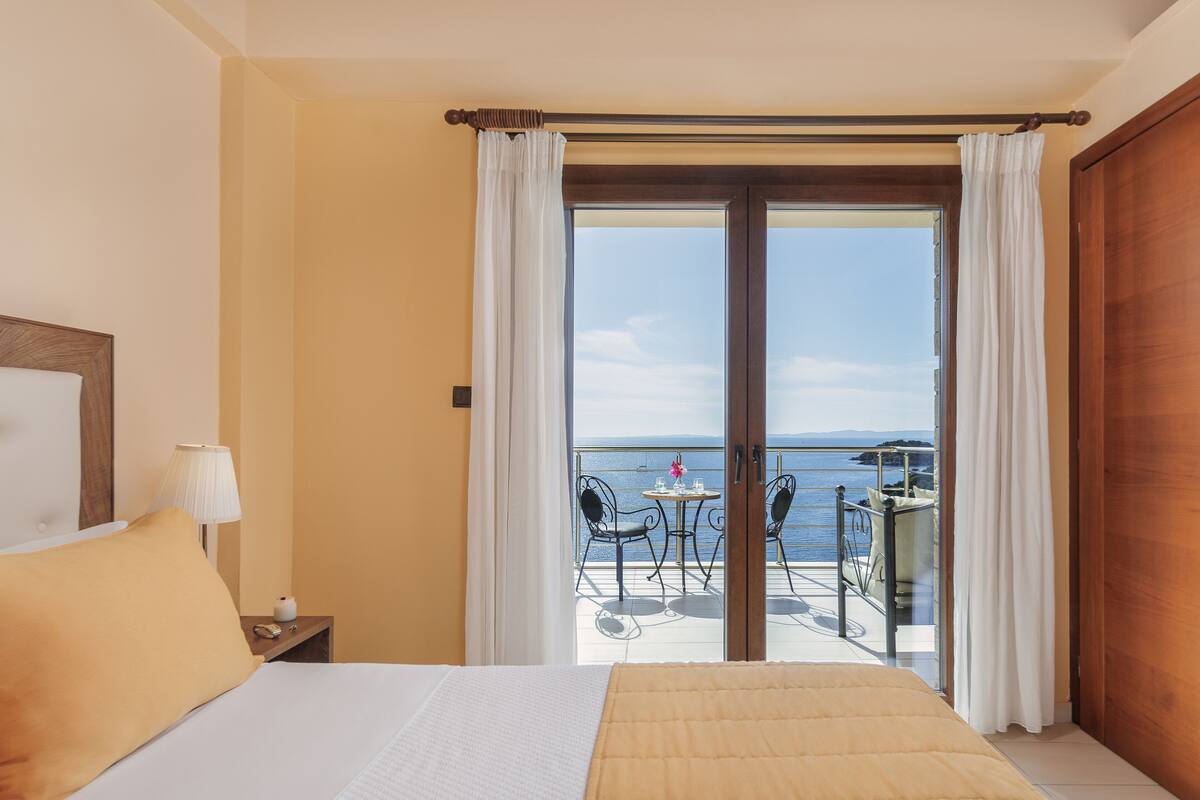 Een hotelkamer met een bed, een nachtkastje, een lamp en uitzicht op een balkon met een tafel en stoelen.