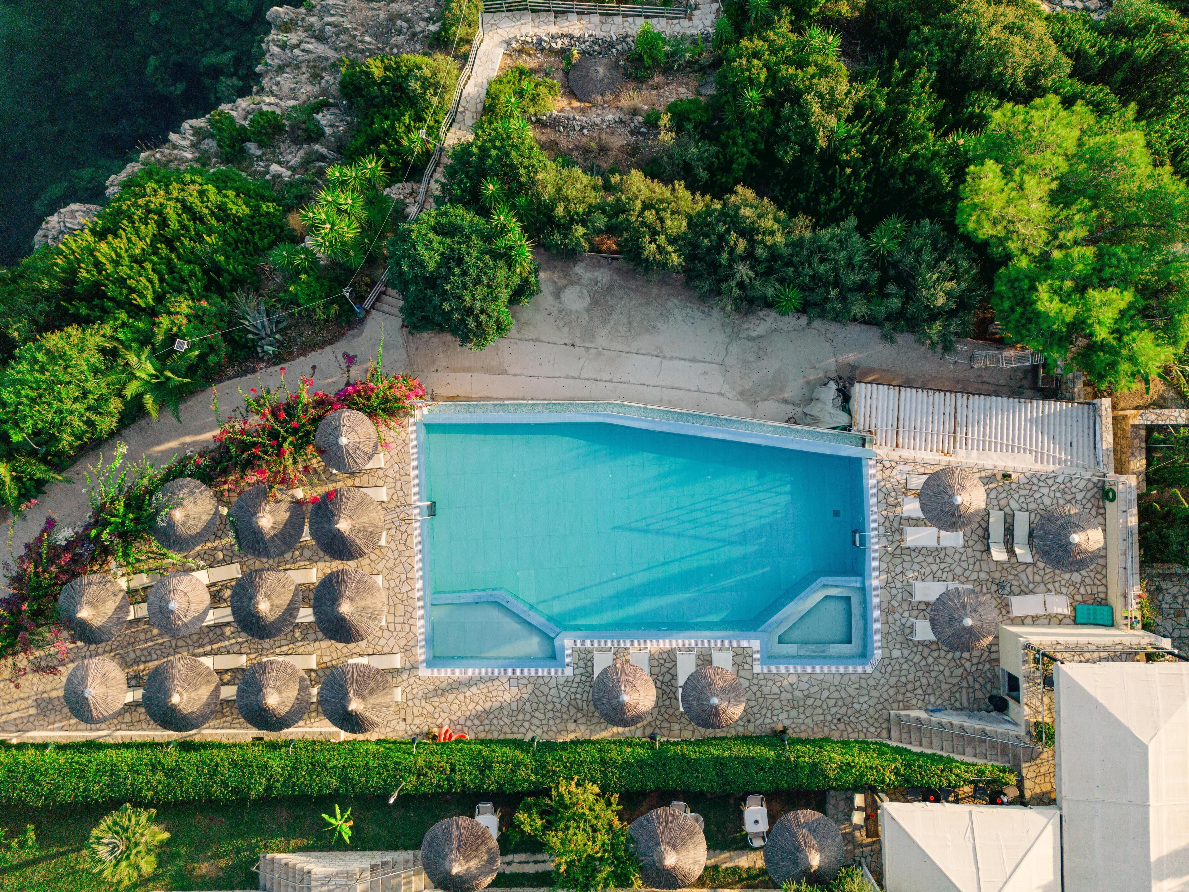 An aerial view of a resort with a large swimming pool surrounded by lounge chairs and umbrellas, set against a backdrop of lush greenery and rocky terrain.