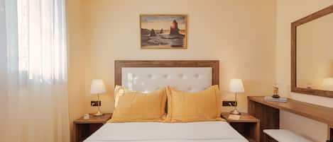 Una habitación de hotel con una cama grande, dos mesitas de noche con lámparas, un banco y un cuadro en la pared, además de un espejo sobre el banco.