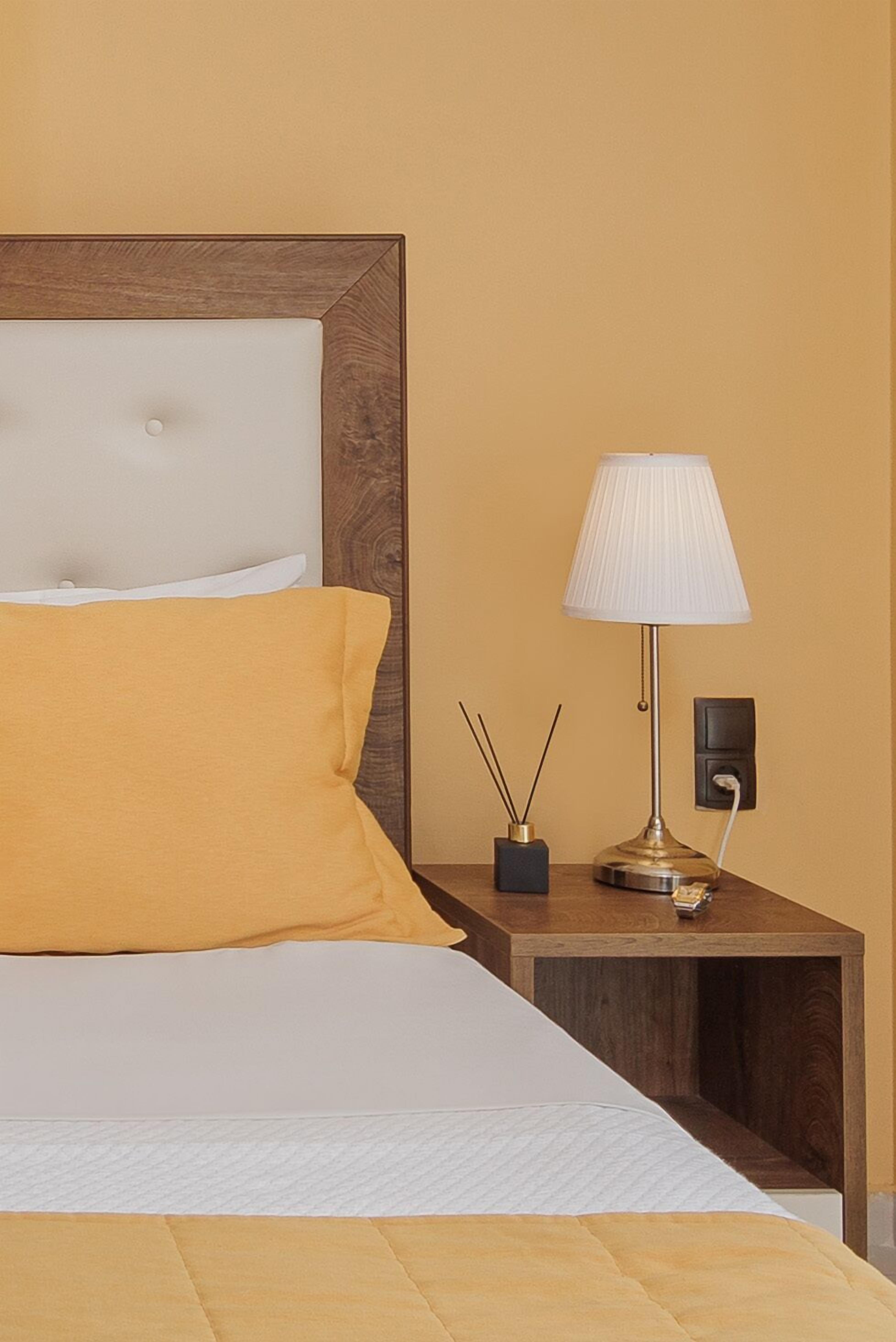 Una cama bien hecha con cabecera blanca y almohada amarilla, una mesita de noche con lámpara, y un pequeño florero con ramas de juncos.