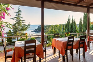 Una terraza con mesas y sillas, con vistas a un paisaje costero con árboles y el mar.
