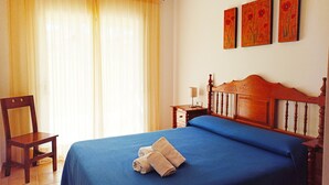 Un lit bien fait, recouvert d’une couverture bleue et agrémenté de deux serviettes, une chaise en bois, une table de chevet avec une lampe et une fenêtre avec des rideaux.