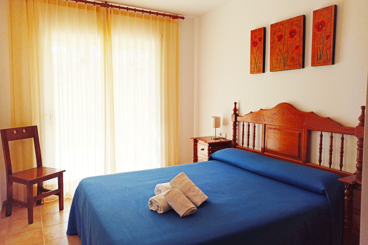 A neatly made bed with a blue bedspread and two towels, a wooden chair, a nightstand with a lamp, and a window with curtains.