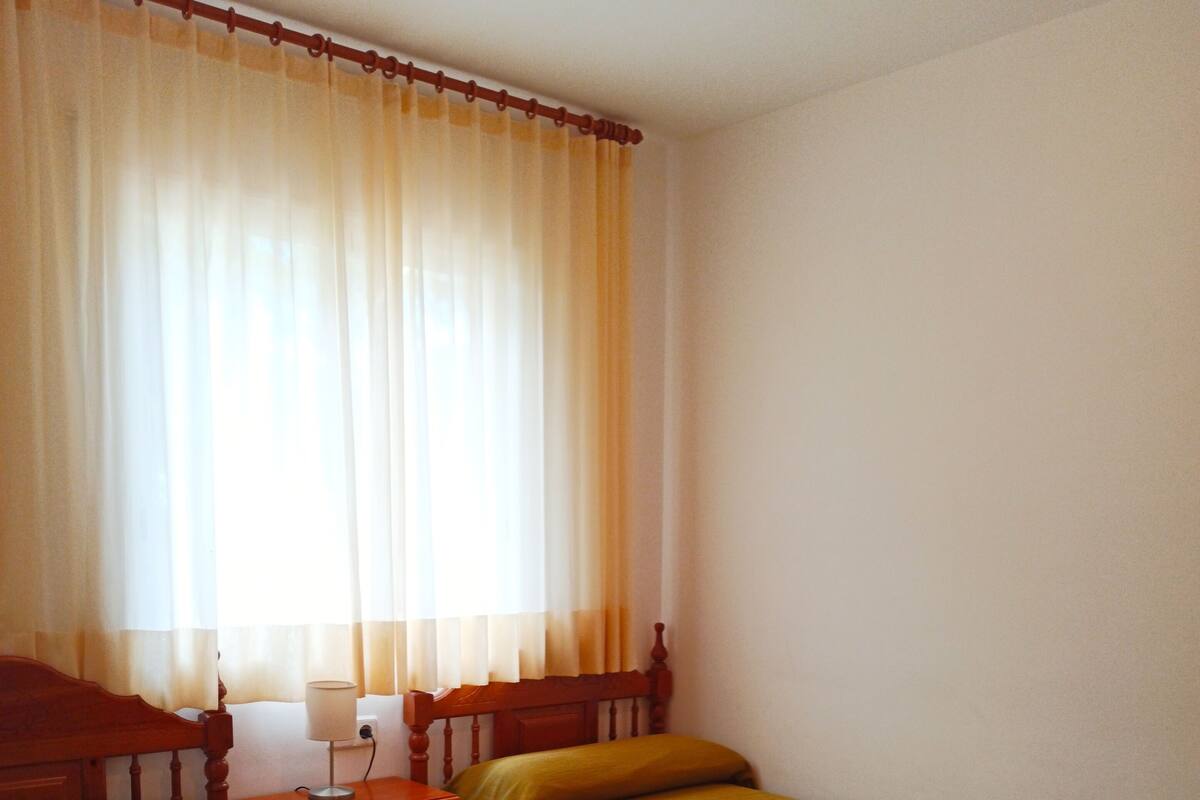 A hotel room with two single beds, a wooden nightstand with a lamp, and a window with curtains.