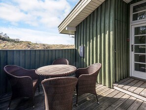 A covered outdoor seating area with wicker chairs and a table, a door with a window, and a green corrugated wall.
