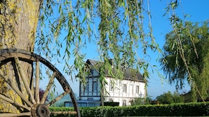 A rustic wooden cart wheel leaning against a tree, a well-maintained lawn, and a large white building in the background. - vacation home   (Hugleville-en-Caux)