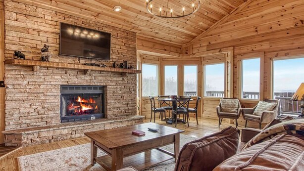 A cozy living room with a fireplace, wooden walls, a large TV, and a balcony with a view. - ⭐️Prime Location with Stunning Views, Hot Tub, Game Room⭐️ (Gatlinburg)
