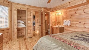 A wooden cabin bedroom with a bed, a ceiling fan, a bathroom with a sink and mirror, and a window with blinds. - ⭐️Prime Location with Stunning Views, Hot Tub, Game Room⭐️ (Gatlinburg)