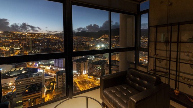 A high-rise room with large windows offering a cityscape view at dusk.