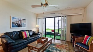 A living room with a black leather sofa, a glass coffee table, a wicker chair, a flat-screen TV, and a large window with a view of the water. - Stunning Bay Views!  Come visit Paradise today! Call today for availability! (Bradenton Beach)