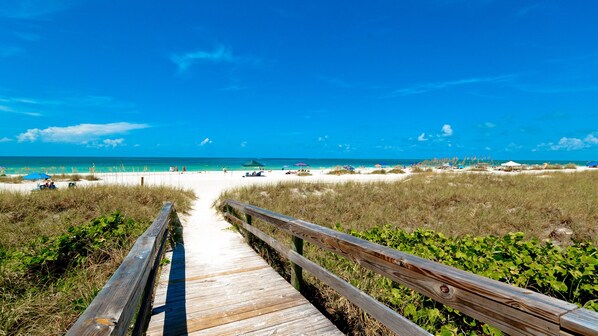 Une promenade en bois menant à une plage où l’eau est d’un bleu limpide et où quelques parasols sont installés.