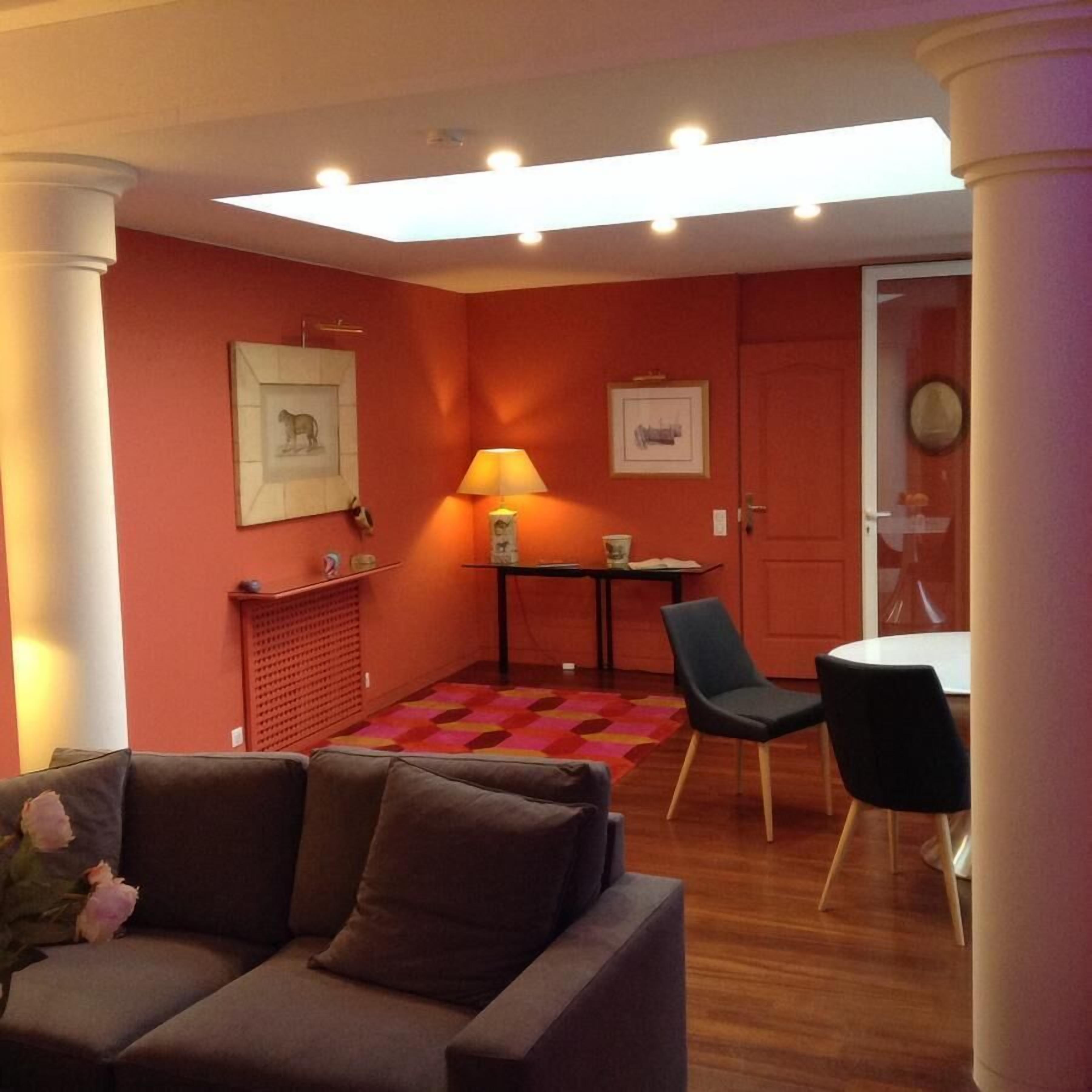 Un salon avec un canapé, une table sur laquelle se trouve une lampe, et un fauteuil. Des tableaux encadrés ornent les murs, et une porte est visible au fond.