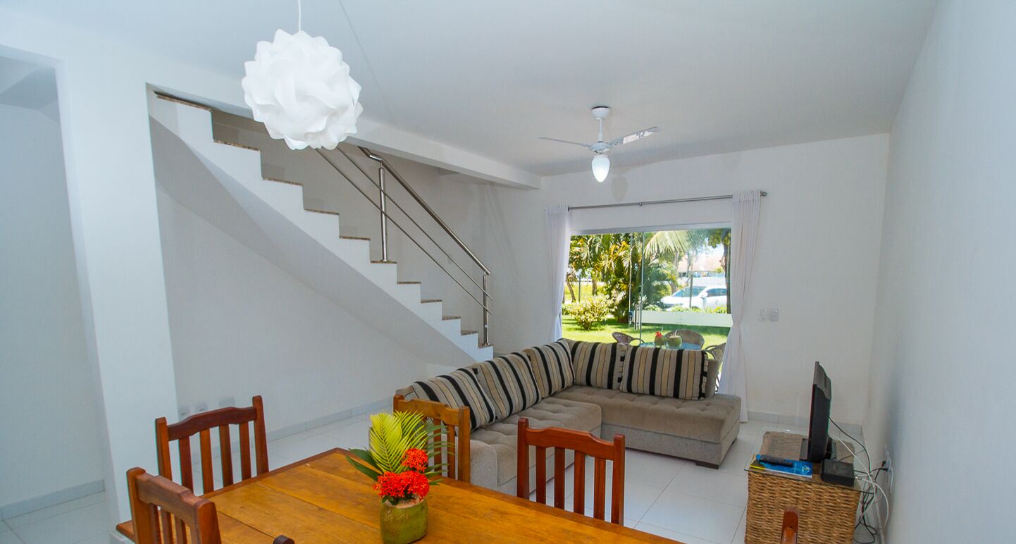 A dining area with a wooden table and chairs, a sofa, a staircase, and a view of greenery outside.