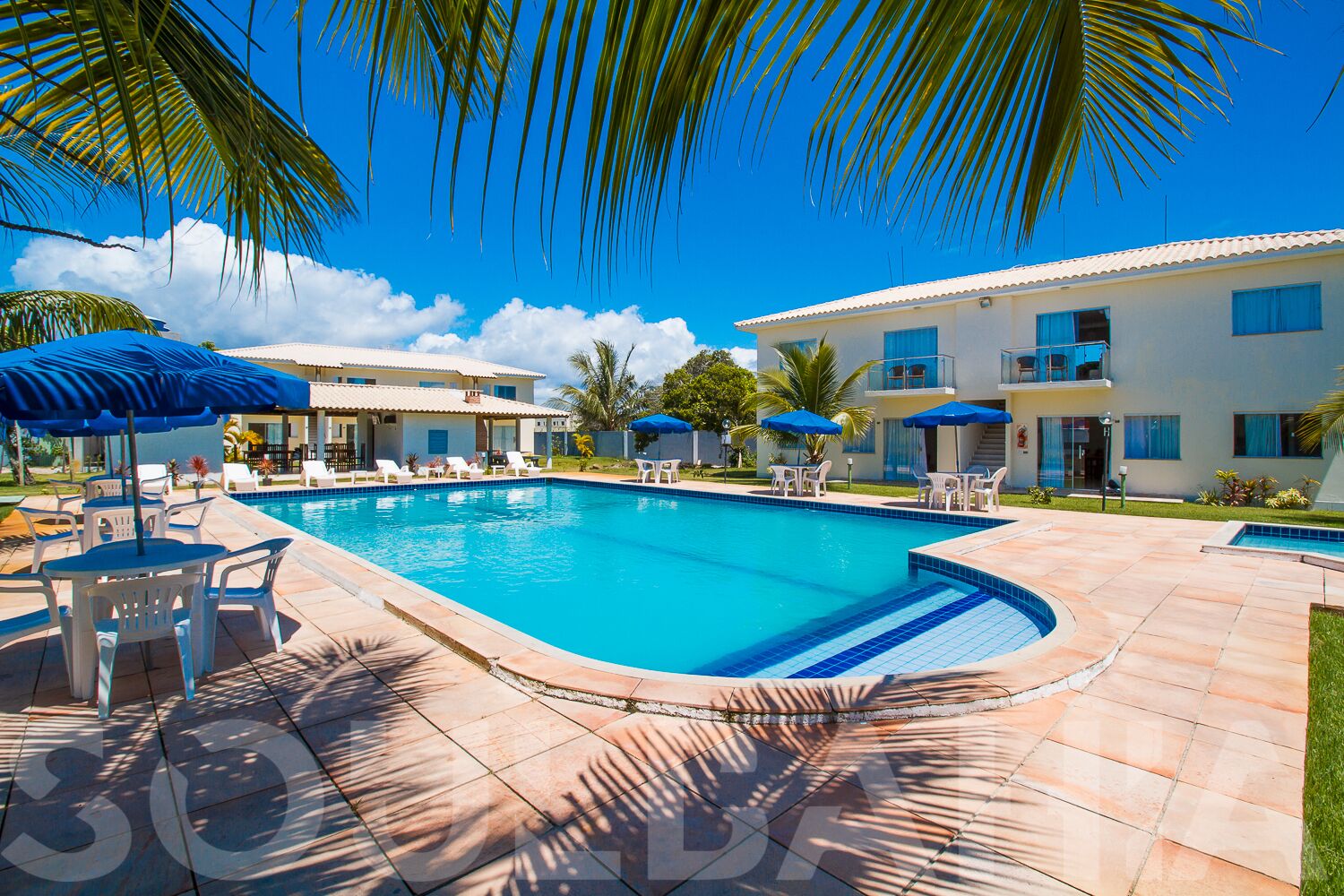 A swimming pool with lounge chairs and umbrellas, surrounded by a building with balconies and a clear blue sky.