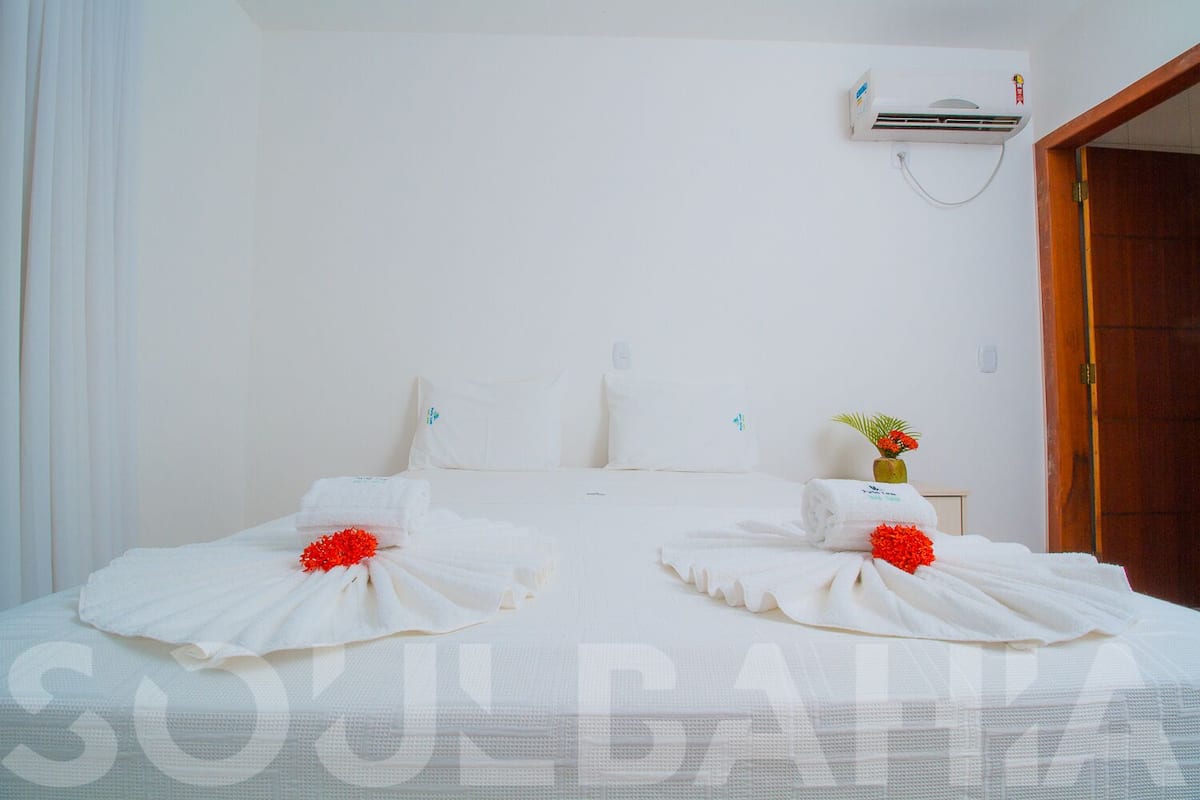 Habitación de hotel con cama, ropa de cama blanca, una mesita con un jarrón de flores y un equipo de aire acondicionado en la pared.