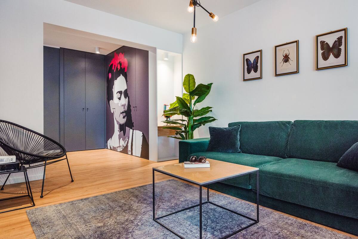 Una sala de estar moderna con un sofá verde, una silla negra, una mesita de centro y una planta grande. En la pared hay tres cuadros enmarcados con mariposas.