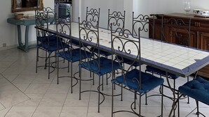 A dining area with a long table, blue upholstered chairs, a white pendant light, and a wooden cabinet. - GITE 10 PERSONNES (Arles)