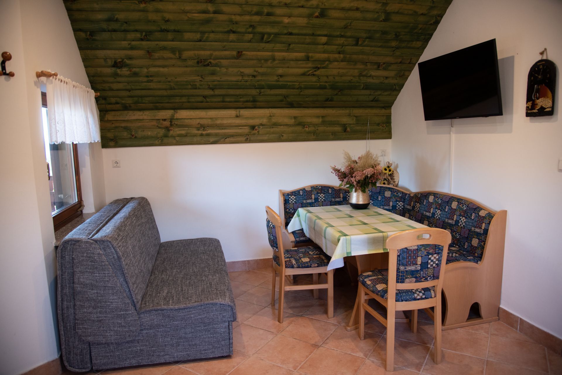 Ein gemütliches Zimmer mit Holzbalkendecke, einer Couch, einem Tisch mit gemustertem Tischtuch und einem an der Wand befestigten Fernseher.