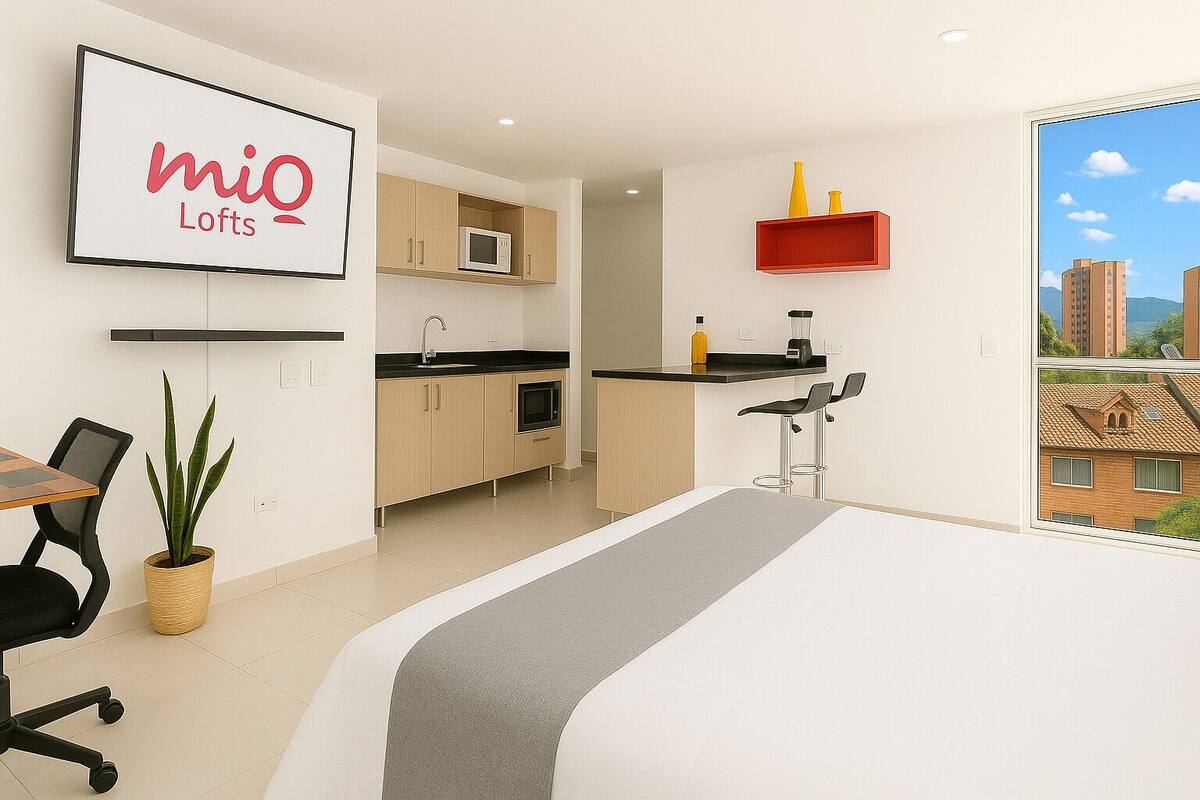 A modern kitchen with a microwave, sink, and bar area. There is a dining table with chairs and a plant. A bed is visible in the foreground with a view of buildings outside.