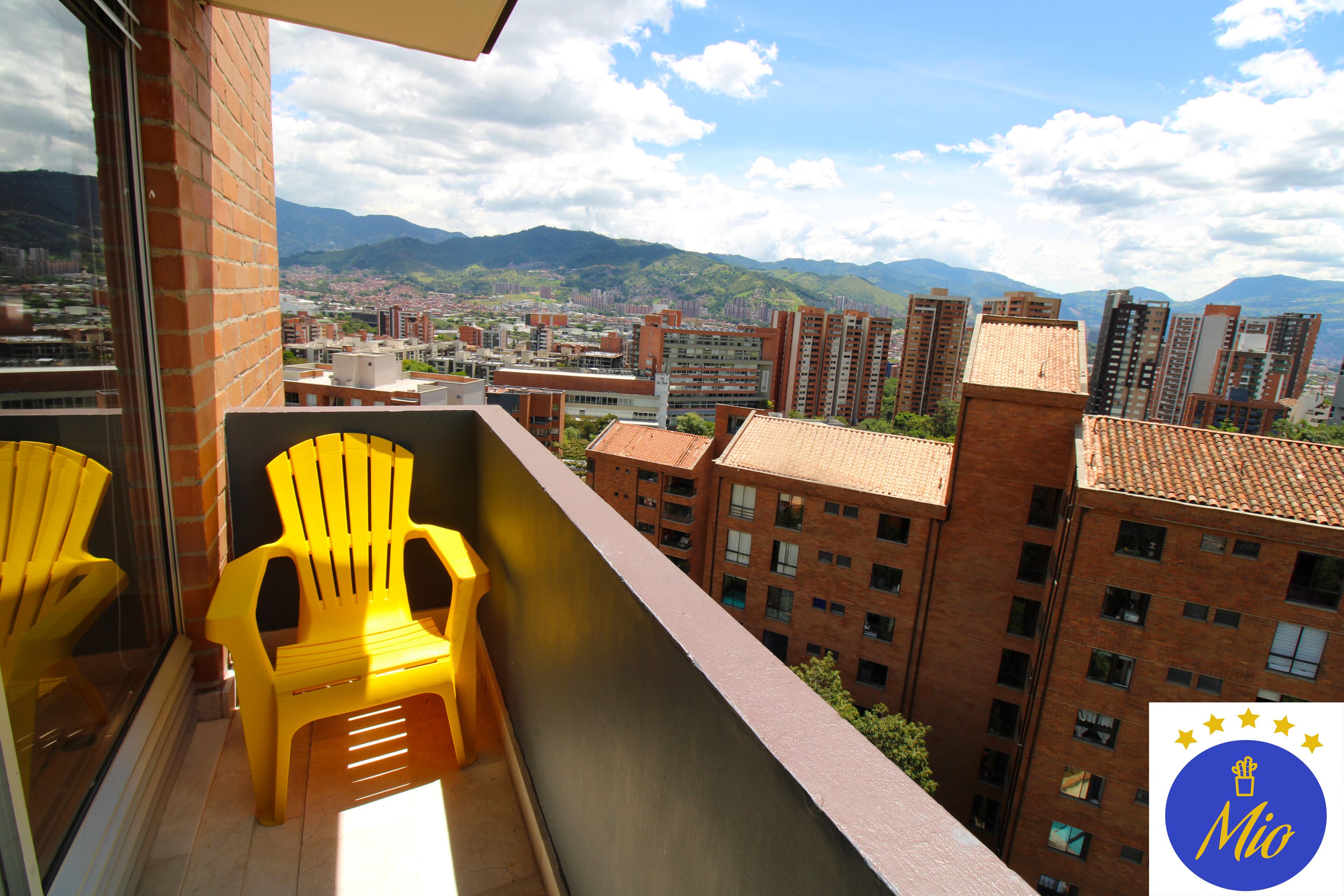 Varanda com cadeiras amarelas, vista para a cidade e montanhas, e um logotipo no canto.