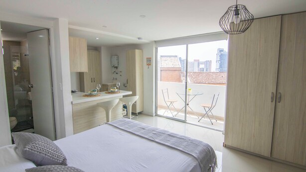 A modern hotel room with a large bed, a bathroom, a kitchenette, and a balcony with a city view. - Mio Lofts (Medellín)