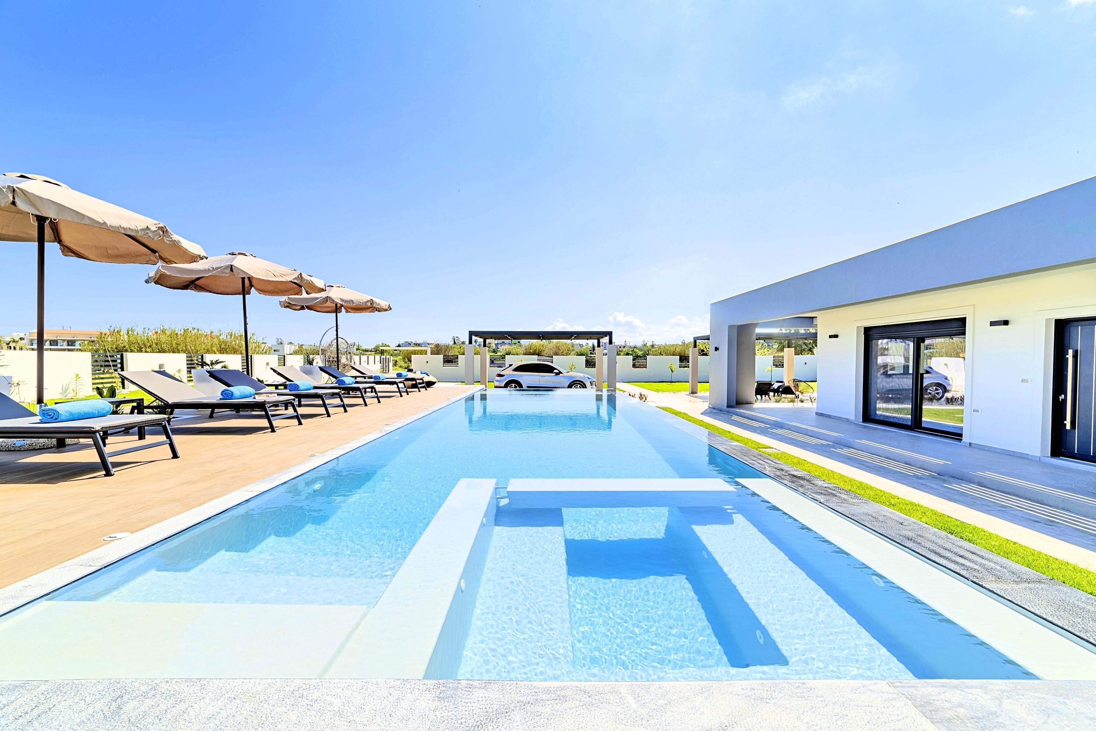 A swimming pool with lounge chairs and umbrellas, a covered area, and a modern building with a car parked outside.