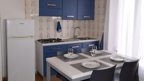 Una cocina compacta con armarios azules, un refrigerador blanco y una mesa de comedor preparada para dos personas, con platos y vasos.