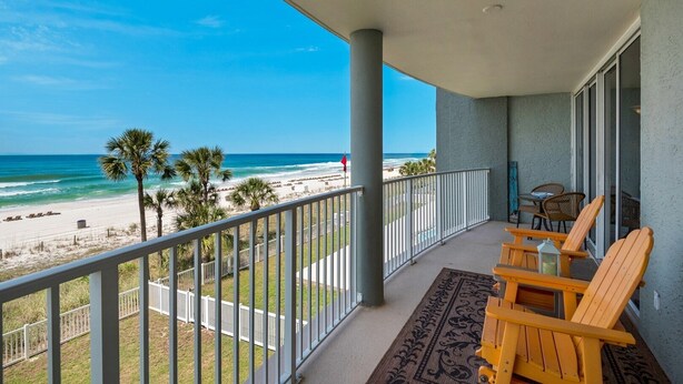 A balcony with a view of the beach and ocean. There are wooden chairs and a table on the balcony.