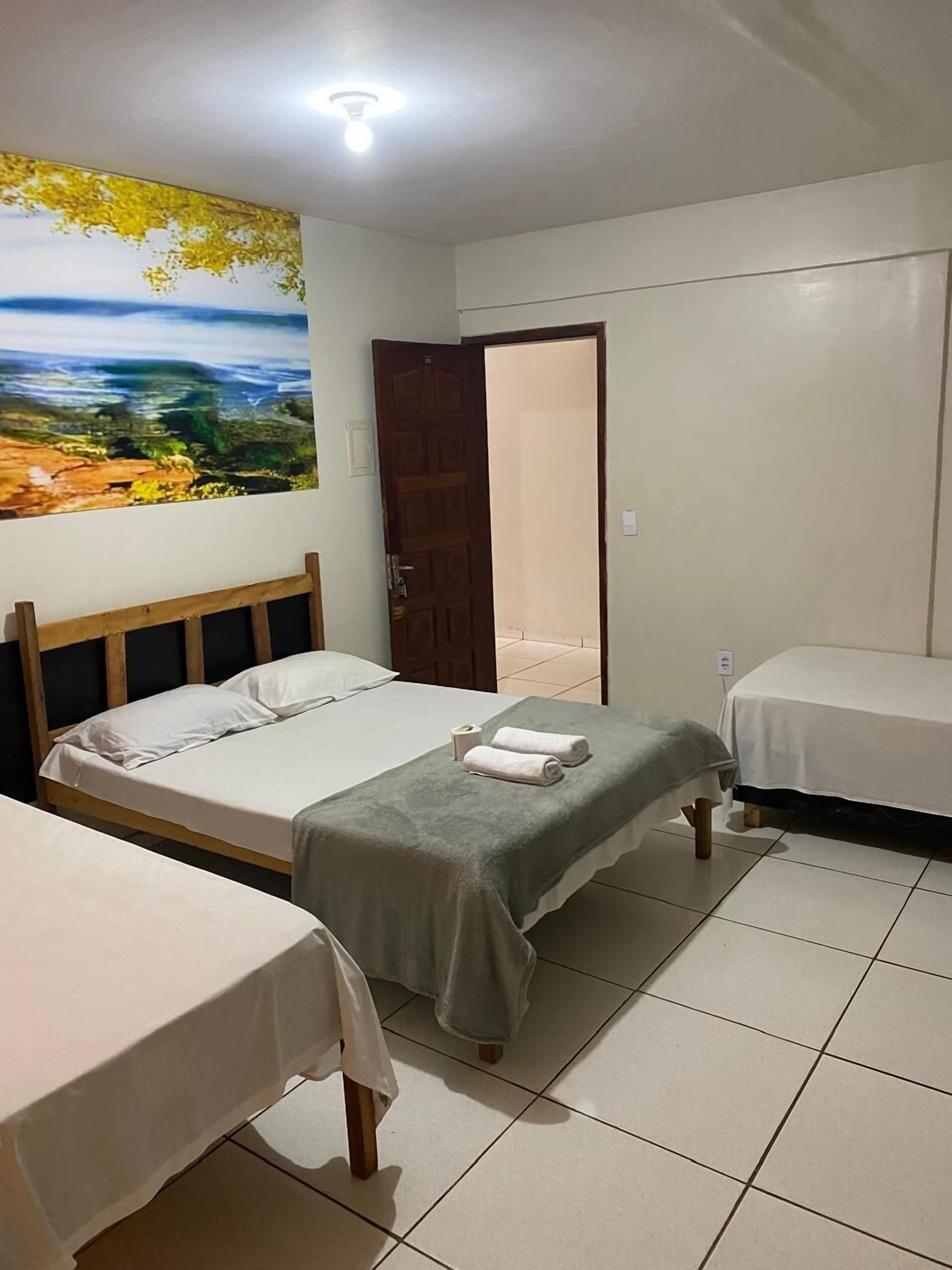 Une chambre avec deux lits, une tête de lit en bois, un tableau représentant un paysage, un sol carrelé et une porte donnant sur une autre pièce.