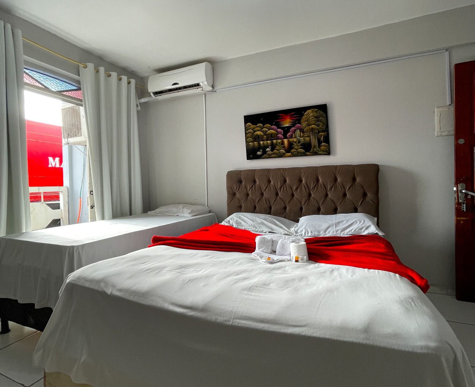 A hotel room with a bed, a window with curtains, an air conditioning unit, and a painting on the wall.