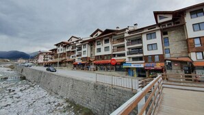 A row of multi-story buildings with balconies, a stone retaining wall, and a wooden boardwalk. - Bansko Royal Towers - Apartments (Bansko)