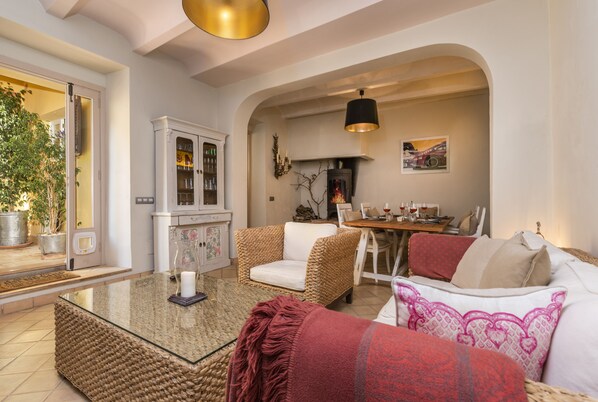 Un salon avec une table basse en verre, des fauteuils en osier, un canapé agrémenté de coussins à motifs, une cheminée et un coin repas avec une table dressée pour deux personnes.
