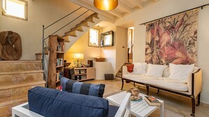 A living room with a blue sofa, white armchair, and a coffee table with books. There is a staircase with a wooden handrail and a large painting on the wall.