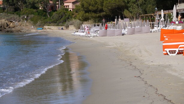 A beach with umbrellas and lounge chairs, a rocky shoreline, and buildings in the background. - NEW: Air-conditioned apartment with wifi (Le Lavandou)