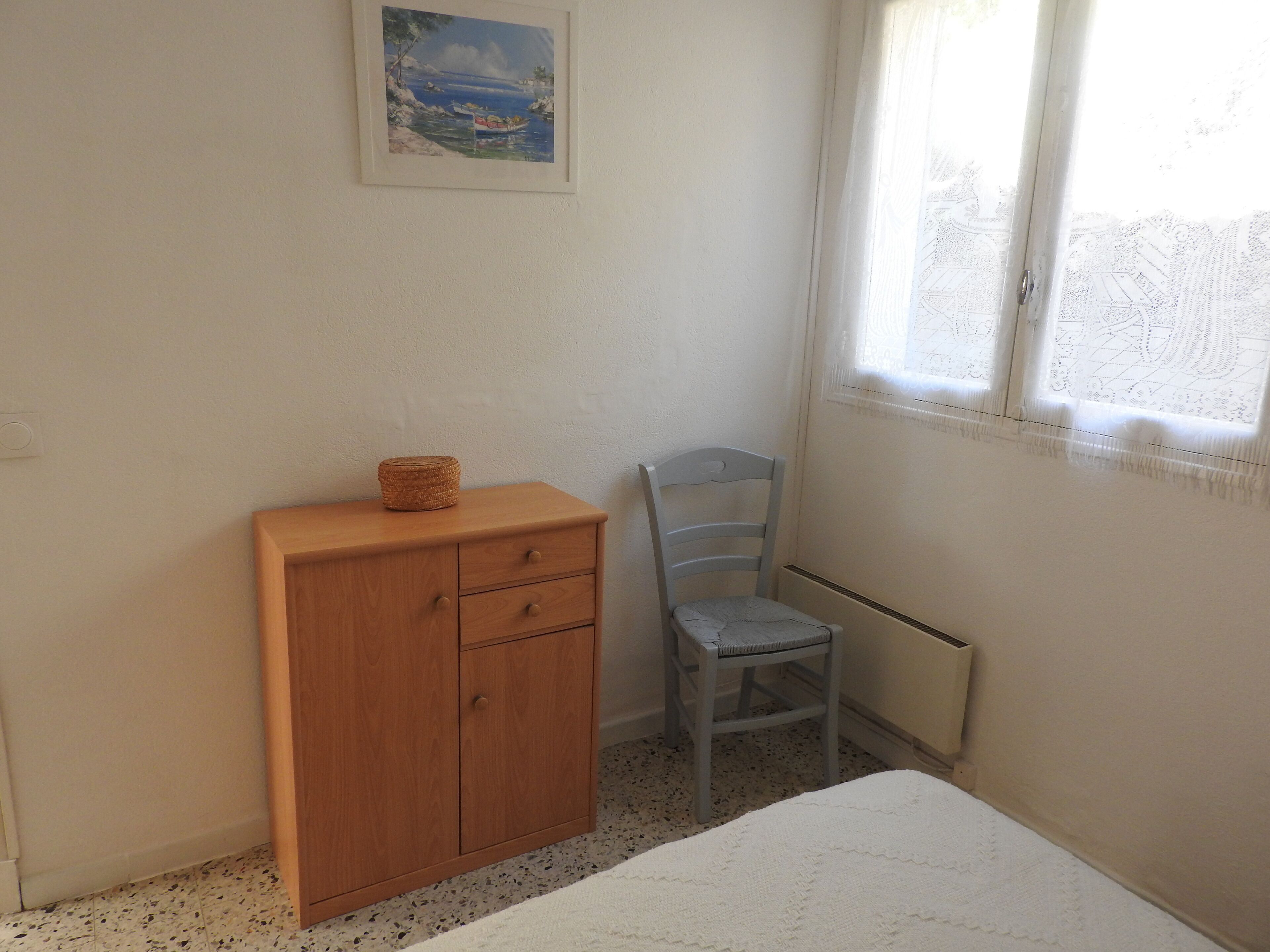 A bedroom with a wooden cabinet, a chair, a bed, and a window with lace curtains.