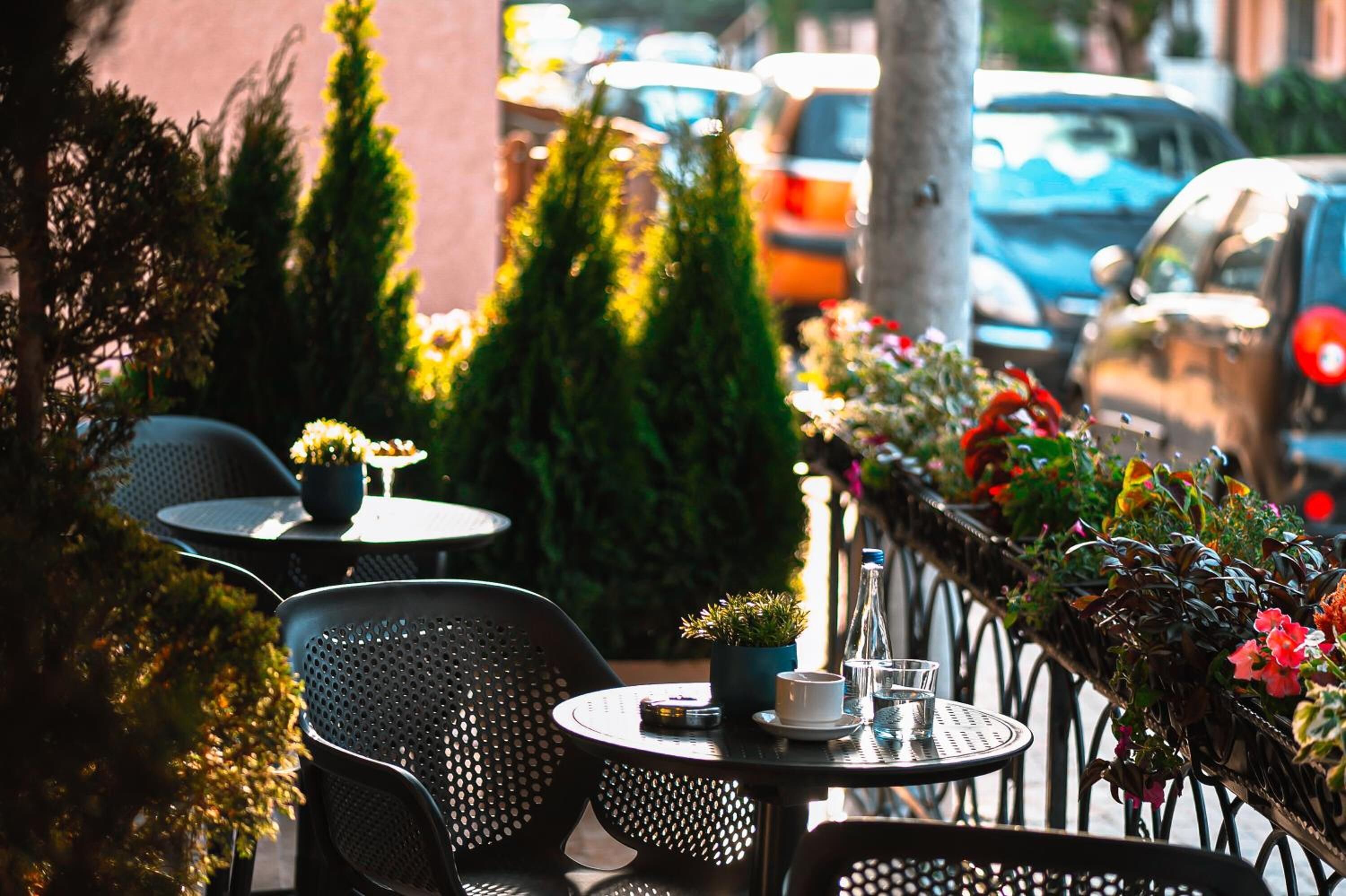 An outdoor cafe with round tables and chairs, potted plants, and a railing with flowers.