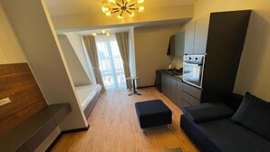 A compact hotel room with a bed, a sofa, a small table, a kitchenette, and a ceiling light fixture. - MM Rooms (Skopje)
