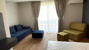 A hotel room with a sofa, ottoman, and armchair, a bed, and a view of the city through large windows. - MM Rooms (Skopje)