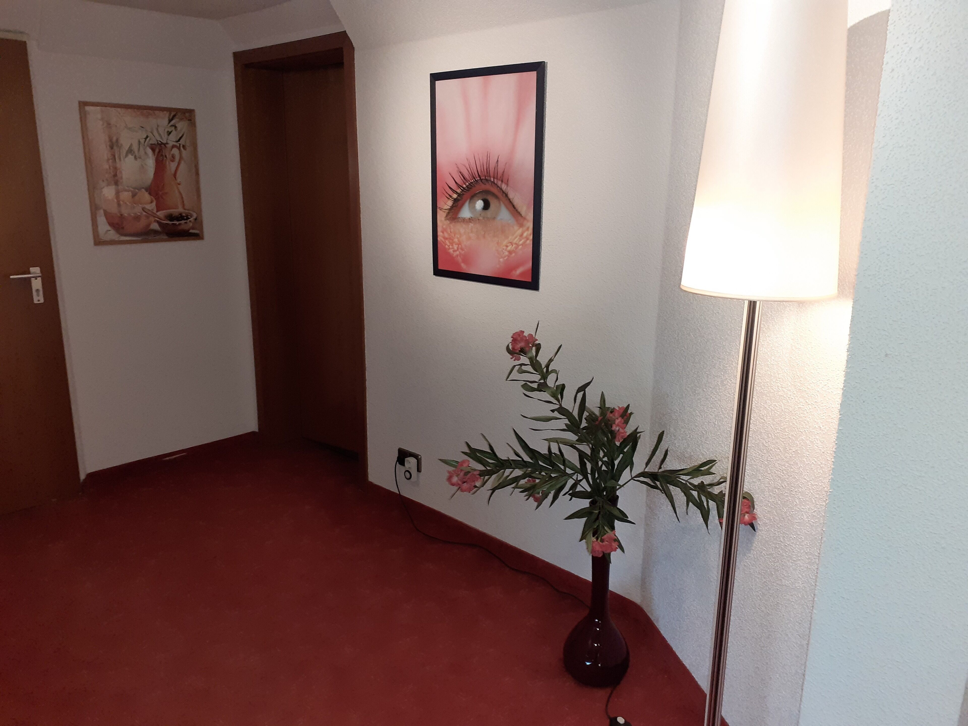A hallway with a red carpet, a vase with flowers, a lamp, and two framed artworks on the wall.