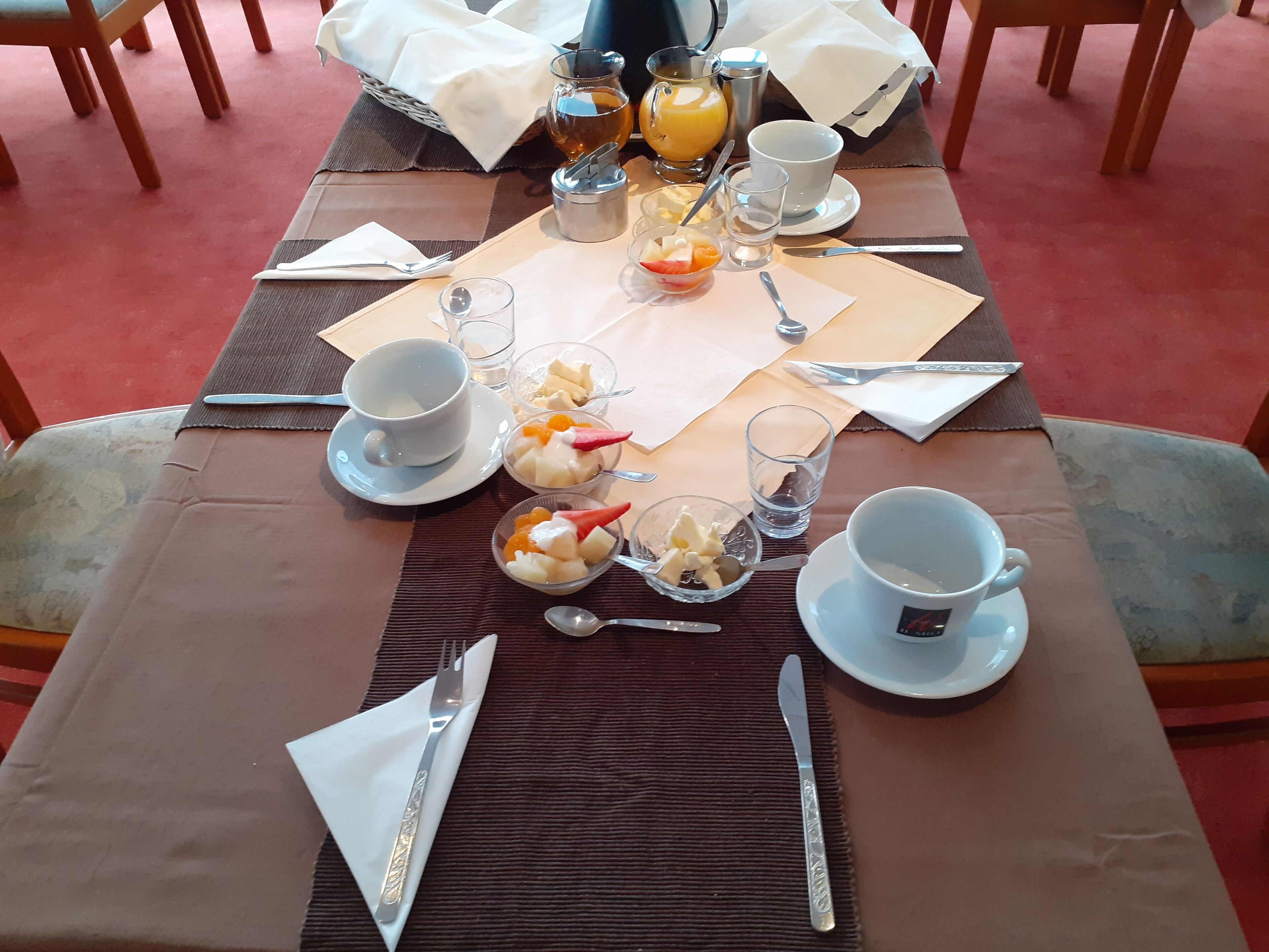 A breakfast table set with a cup, a glass, a bowl of fruit, a glass of juice, a small pitcher, and a knife and spoon.