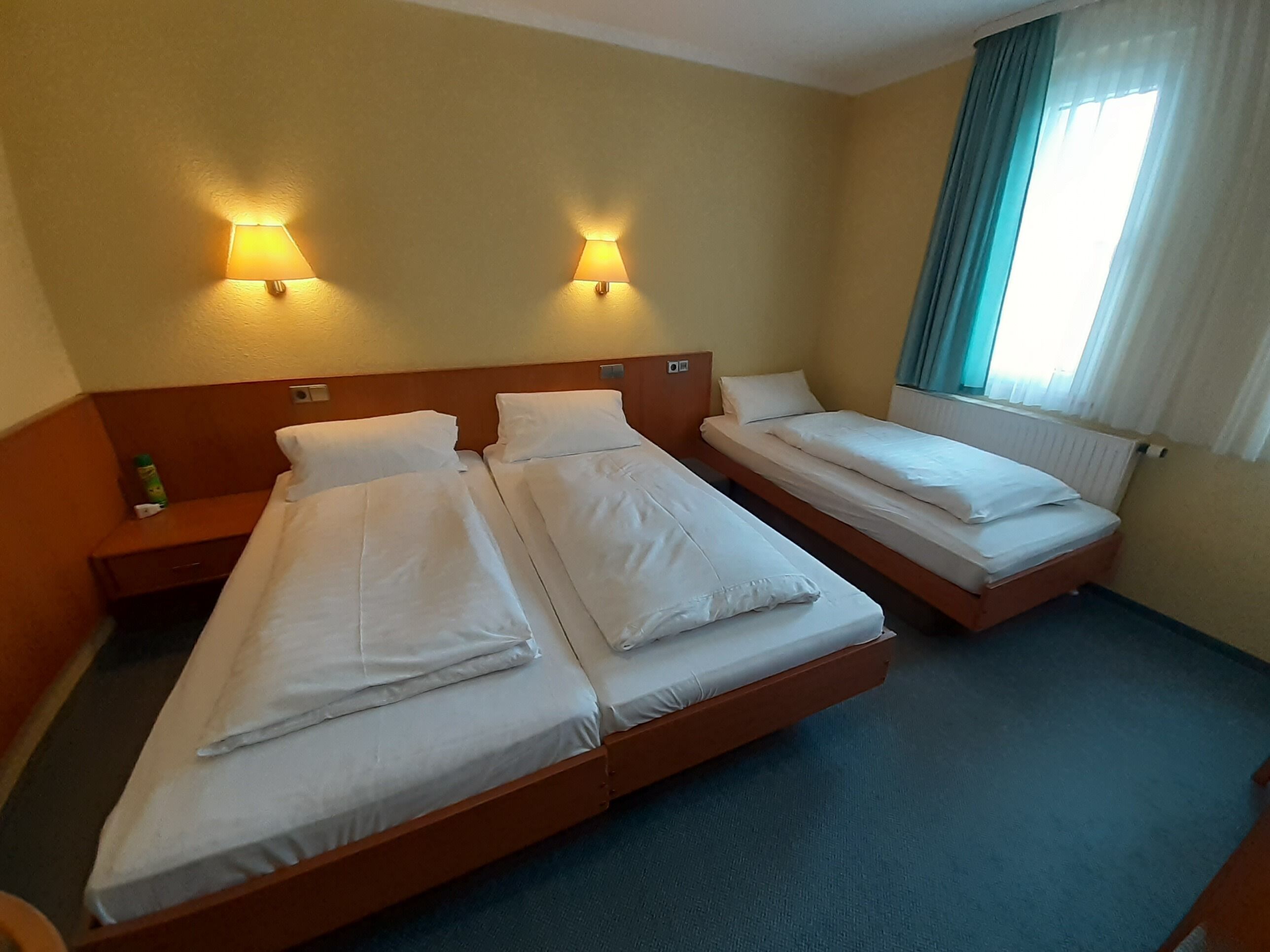 A hotel room with two single beds, a wooden headboard, a nightstand with a lamp, and a window with curtains.