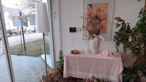 A room with a large window, a table with a pink tablecloth, a vase with dried flowers, and a wicker basket with candles. - Edit Pension Rossau (Rossau)