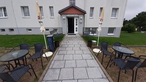 A hotel entrance with a paved walkway leading to a seating area with tables and chairs. - Edit Pension Rossau (Rossau)