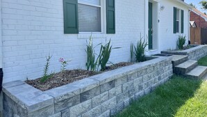 A white brick house with green shutters, a stone retaining wall, and a small garden with plants and a pink flower. - Newly Remodeled! Near Old Town - 2 metros - Solar Home away from home 1600sq (Alexandria)