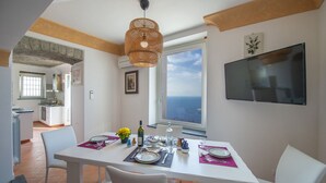 Ein Essbereich mit einem weißen Tisch, gedeckt für vier Personen, einem großen Fenster mit Meerblick und einem an der Wand befestigten Fernseher.