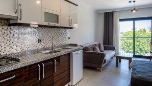 A modern kitchen with a tiled backsplash, stainless steel appliances, and wooden cabinets. There is a dining area with a sofa and a balcony with a view.